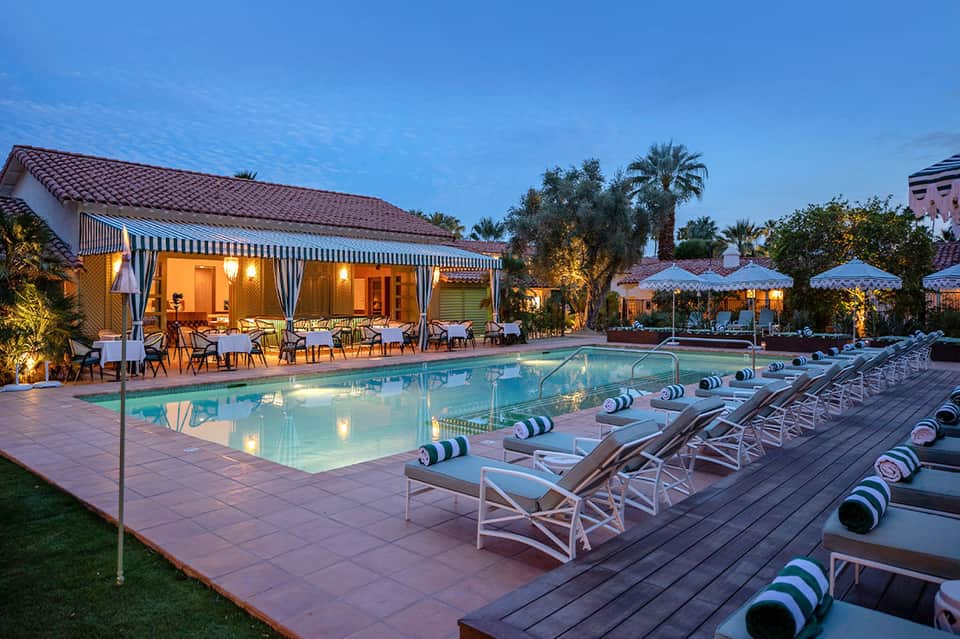 Resort pool at evening with striped loungers, umbrellas, lit clubhouse, and palm trees