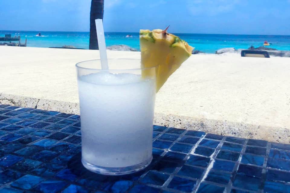 Tropical cocktail with pineapple garnish on beach table with ocean and boats in background