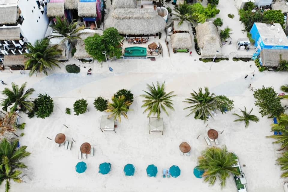 Aerial view of beachfront resort with white sand, umbrellas, tiki huts, palm trees, and turquoise ocean