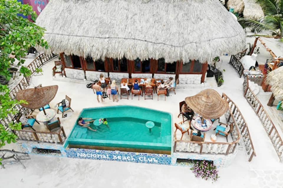 Aerial view of beachfront resort with thatched-roof bar, turquoise pool, and white sand