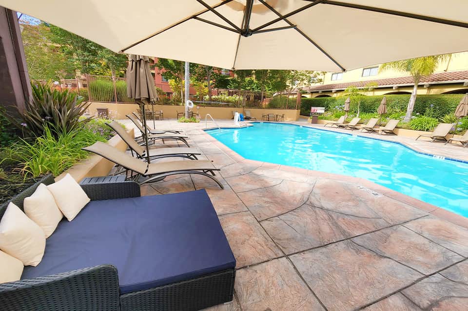Resort pool with lounge chairs, umbrella, natural landscaping, and covered pergola structure