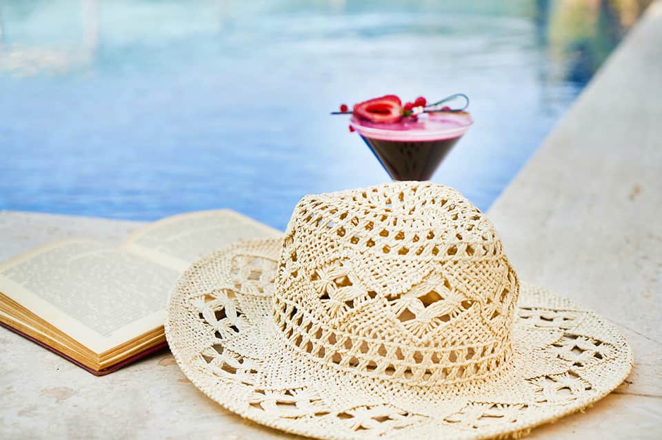 Straw hat and open book by pool with colorful cocktail and ocean view in background