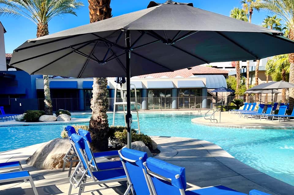 Resort pool area with large black umbrella, blue lounge chairs, palm trees, and modern buildings