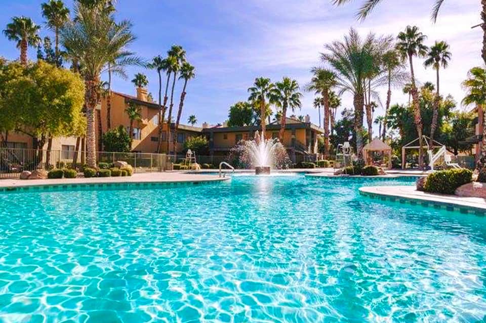 Large resort pool with fountain, palm trees, loungers, and Spanish-style buildings