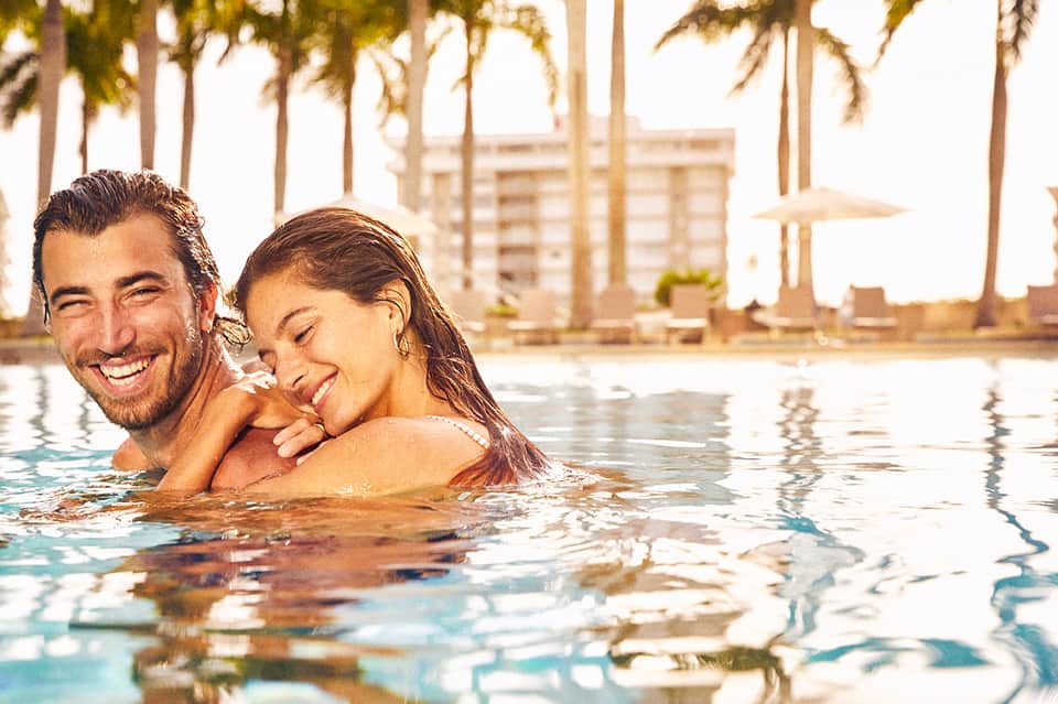 Couple enjoying tropical pool with palm trees and resort buildings at sunset