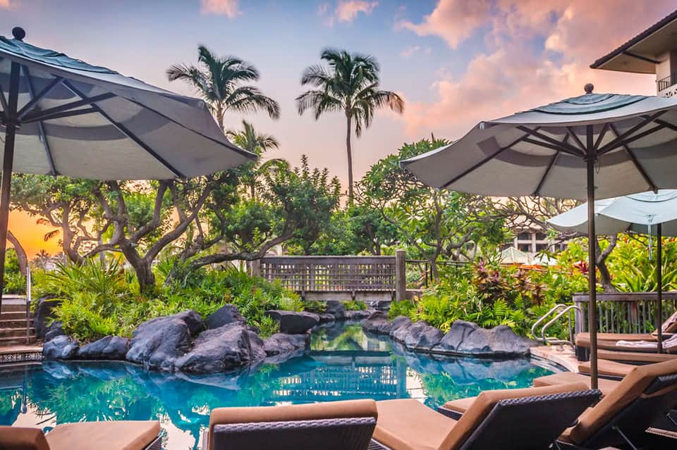 Luxury resort pool with turquoise water, rocky features, palm trees, and lounge chairs under umbrellas at sunset