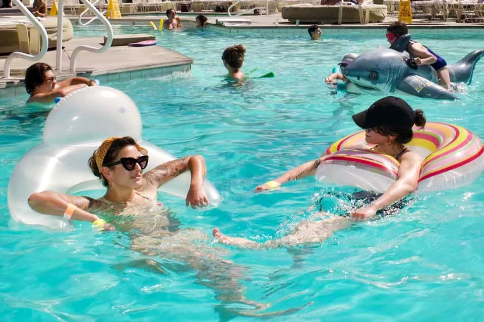 Resort pool with guests swimming, inflatable floats, and lounge seating