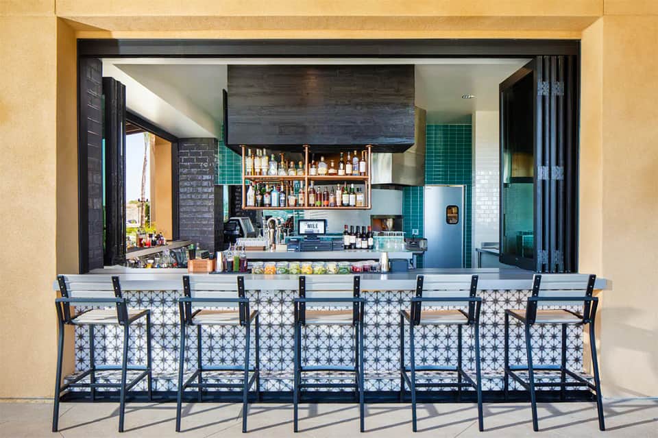 Outdoor bar with blue patterned bar stools facing indoor kitchen with green tile accents