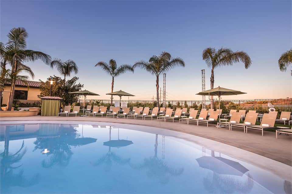 Resort pool at dusk with palm trees, lounge chairs, umbrellas, and amusement park rides visible
