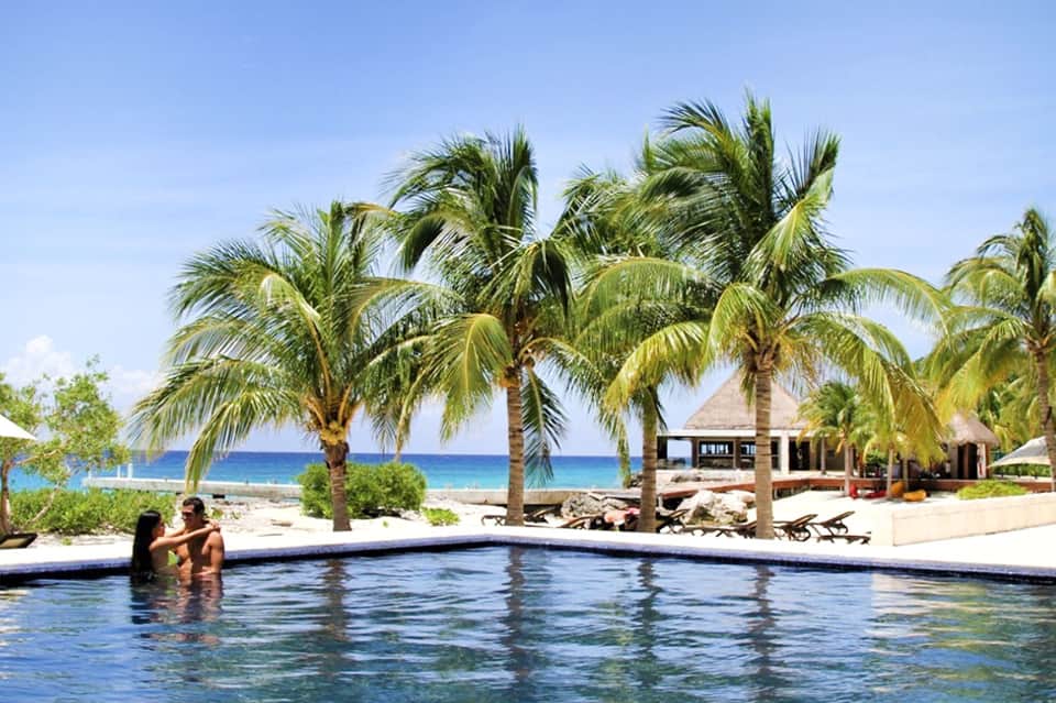 Couple wading in a tropical resort infinity pool with palm trees and beach bungalows visible