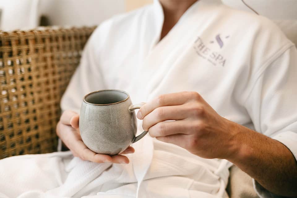 Person in white spa robe holding ceramic mug for hot beverage at wellness resort