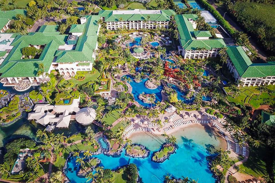 Aerial view of large resort with multiple pools, lush landscaping, and surrounding guest buildings with green roofs