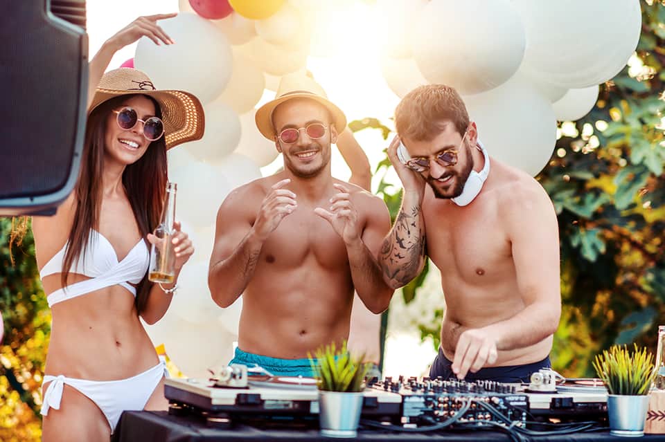Three people at an outdoor DJ booth with white balloons and tropical foliage