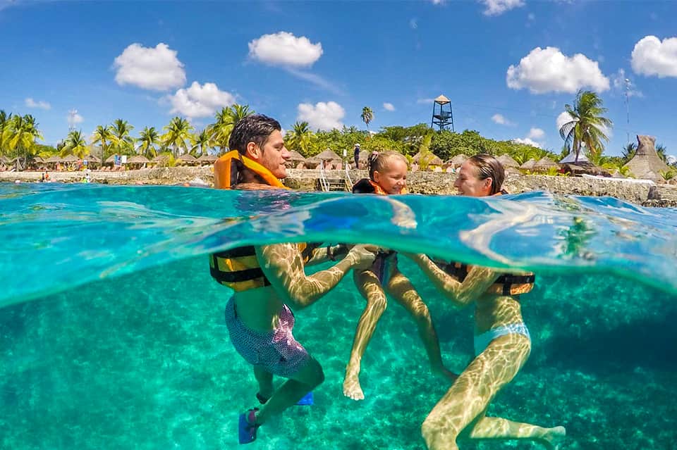 Family snorkeling in crystal-clear turquoise water with tropical beach and palm trees