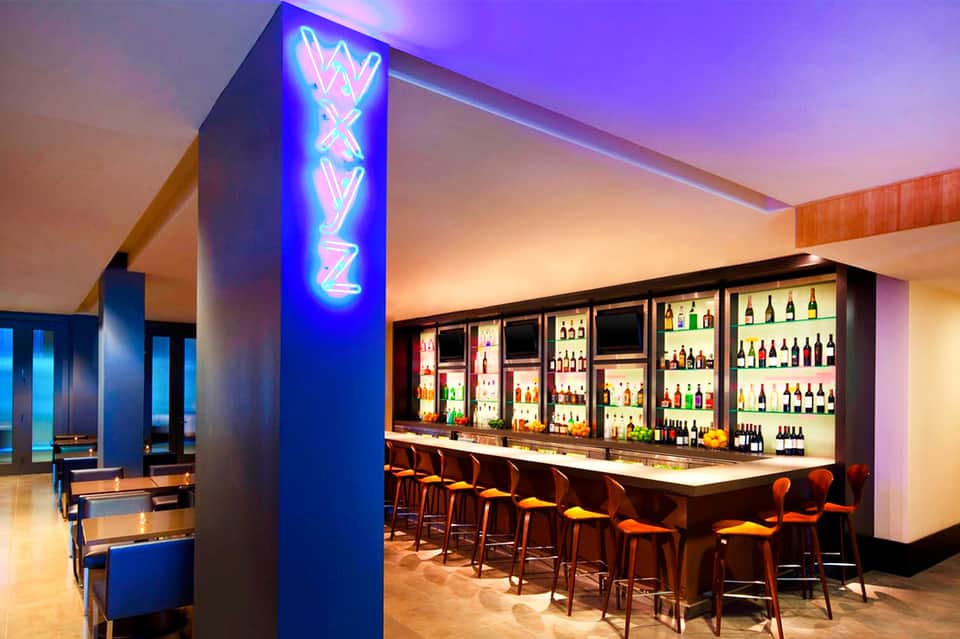Modern lounge bar with neon signage, illuminated liquor shelves, and ambient purple lighting