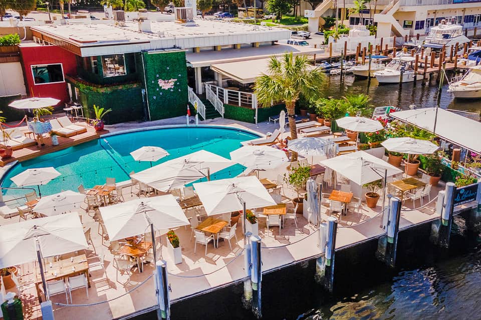 Aerial view of resort pool surrounded by white umbrellas, wooden loungers, waterfront docks, and boats.