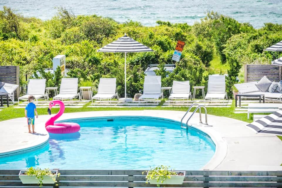 Resort swimming pool with pink flamingo float, white lounge chairs, striped umbrellas, and ocean view beyond vegetation