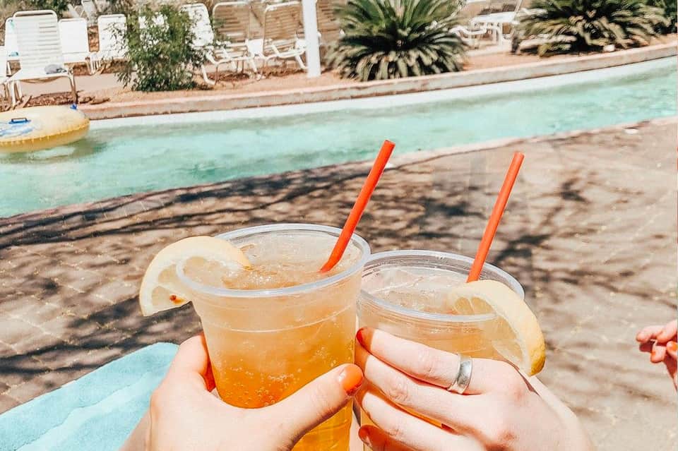 Two tropical cocktails with orange straws by a turquoise pool with palm trees and lounge chairs