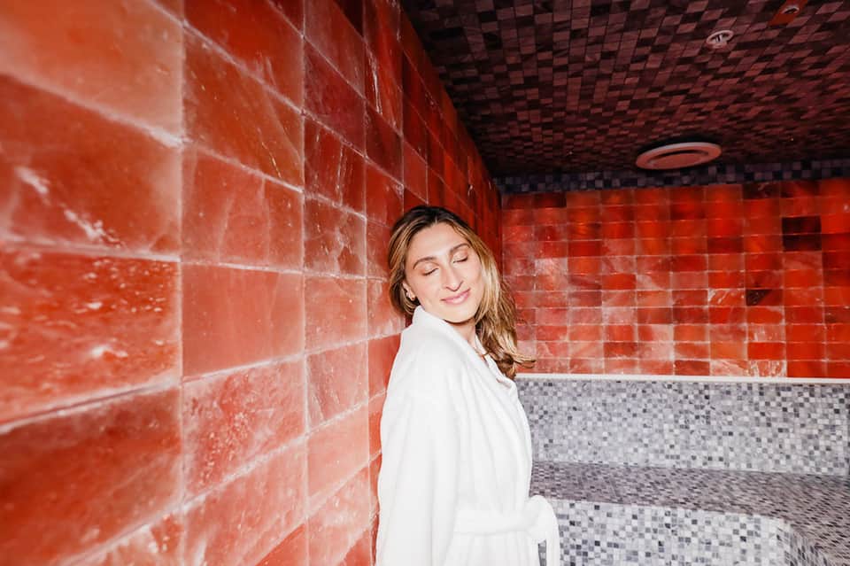 Woman in white robe standing in spa with red tile walls and mosaic floor accents