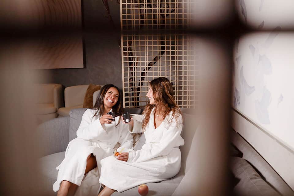 Two women in white robes enjoying beverages in a modern spa relaxation room
