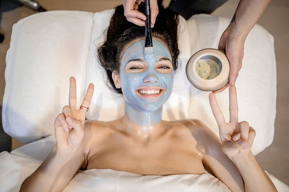 Woman receiving blue facial mask treatment at spa with professional makeup artist