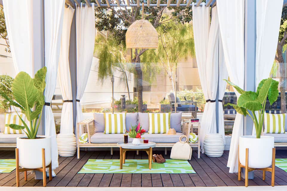 Outdoor cabana lounge with white curtains, striped cushions, woven pendant light, and tropical greenery