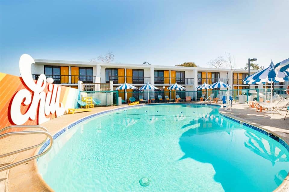 Vintage-style motel pool with retro sign, striped umbrellas, colorful chairs, and mid-century architecture