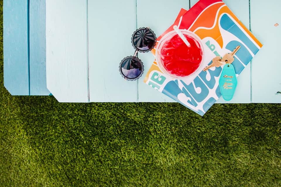 Overhead view of sunglasses, beach towel, sunscreen bottle, and tropical drink on grass