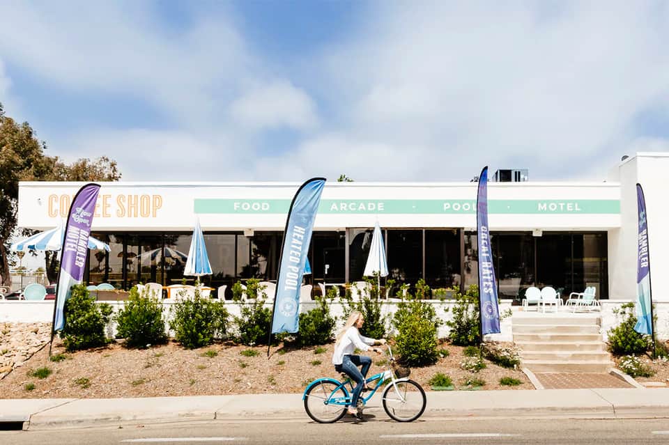 Modern beachside building with coffee shop, food, arcade, pool, and motel amenities; person riding blue bicycle in front