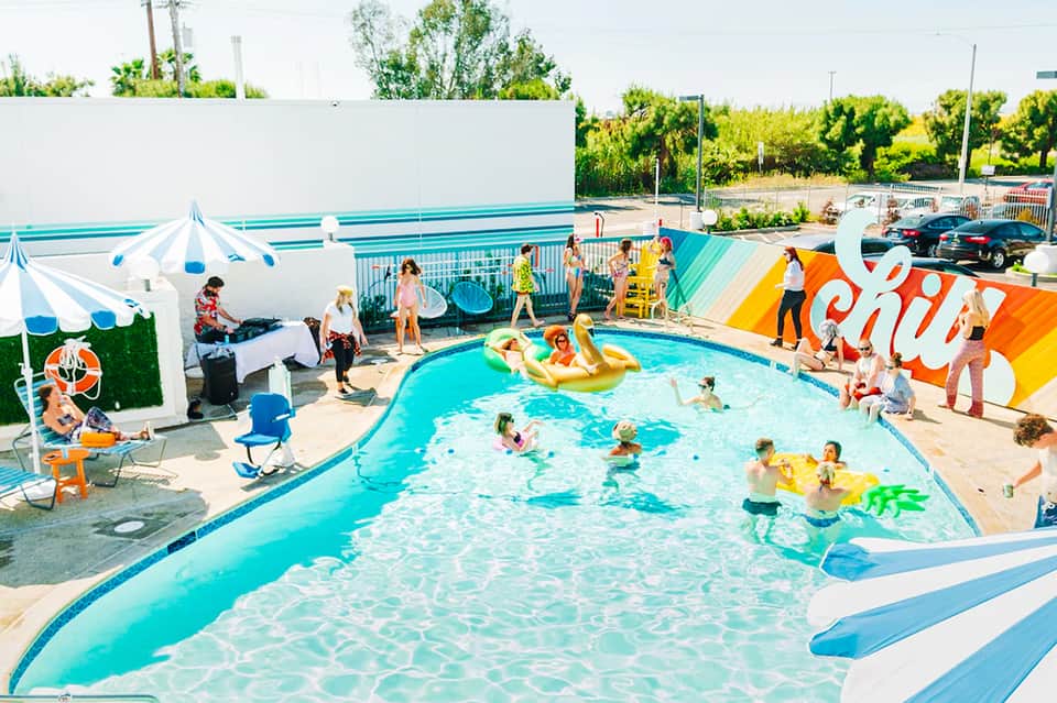 Colorful community pool party with children swimming, inflatable toys, staff in yellow shirts, and striped umbrellas