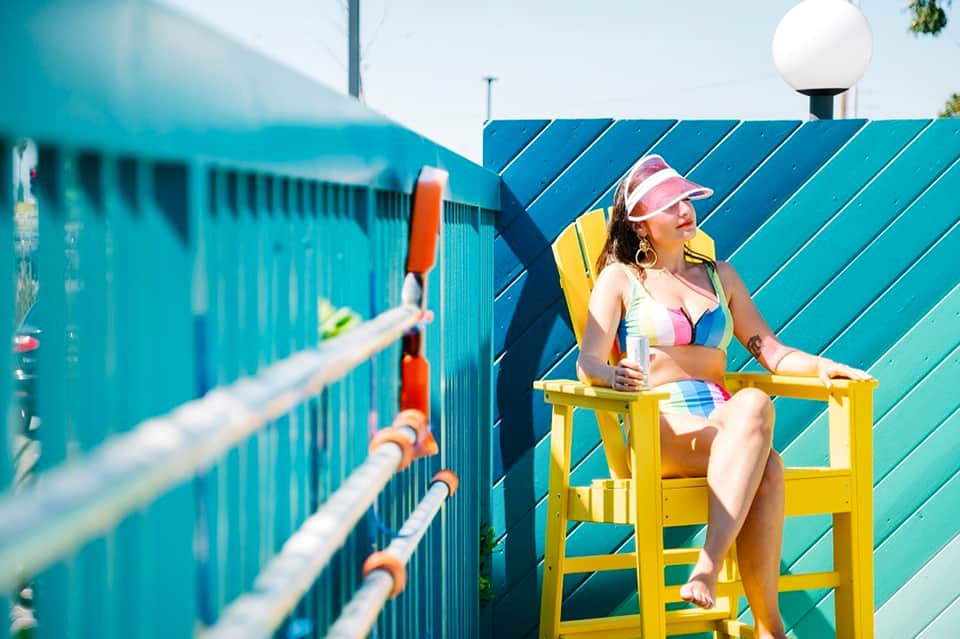 Woman in colorful bikini sitting on yellow lifeguard chair by turquoise pool fence with buildings