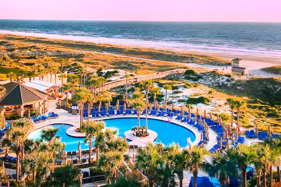 Aerial view of beachfront resort with curved blue pool, palm trees, and sandy beach at golden hour