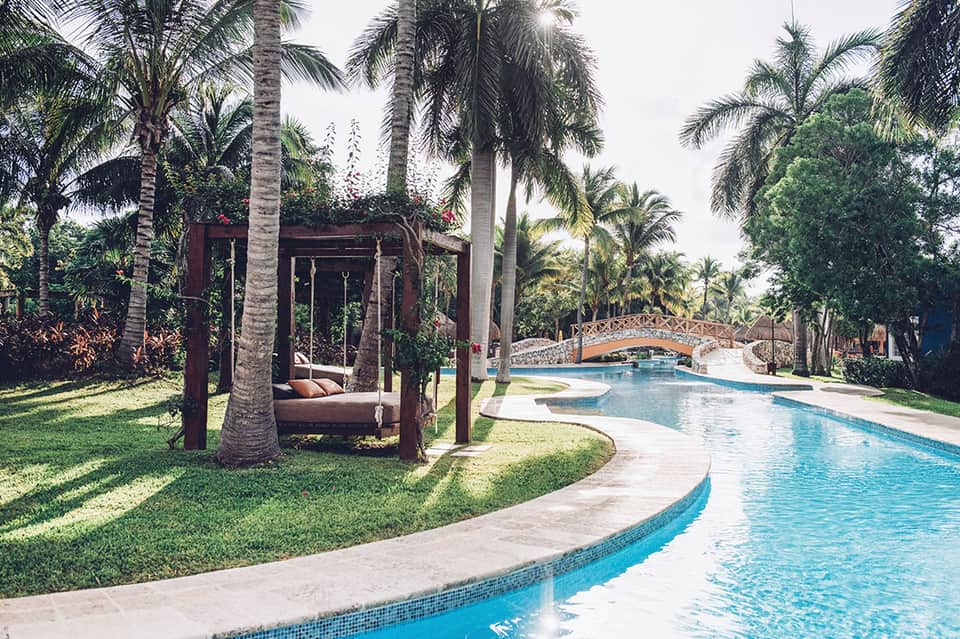 Tropical resort grounds with curved swimming pool, palm trees, wooden pergola structure, and decorative bridge