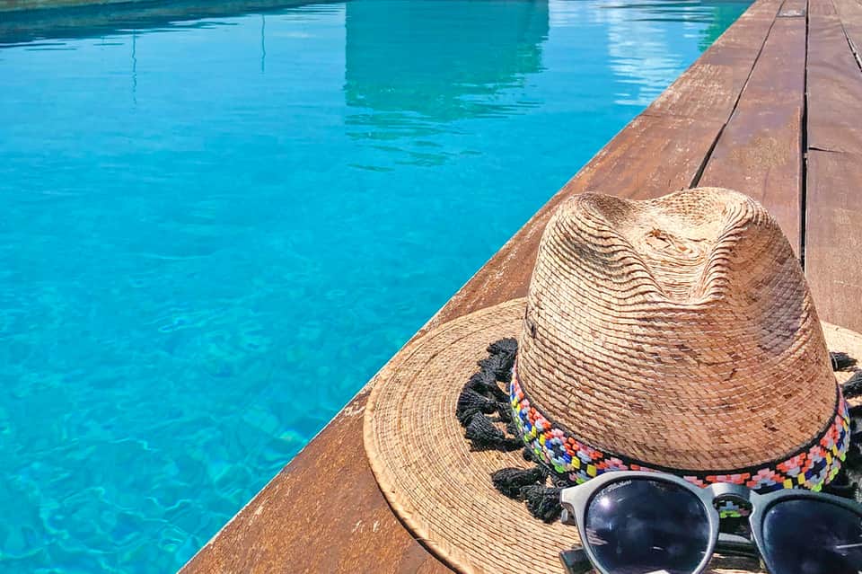 Straw hat and sunglasses resting on wooden deck beside turquoise swimming pool