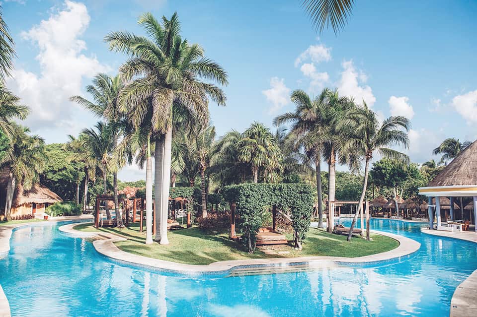 Resort swimming pool with curved design, palm trees, tiki huts, and manicured lawn area