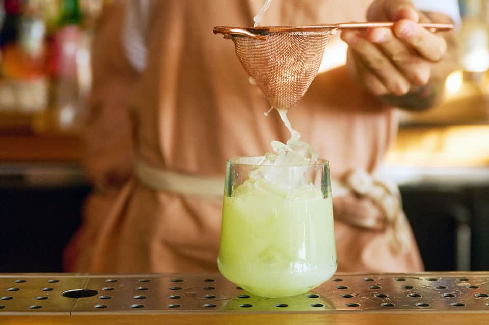 Bartender straining yellow-green cocktail into glass at bar