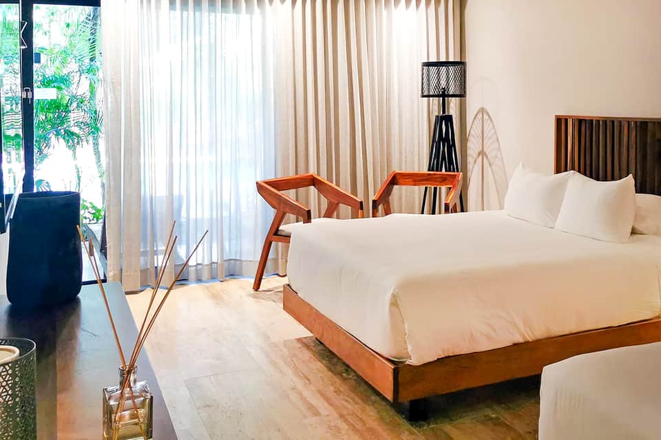 Bright hotel bedroom with white bedding, modern wood furniture, and balcony overlooking tropical landscape