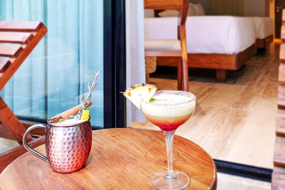 Tropical resort room with cocktails on wooden table, bed visible, and ocean view through glass doors
