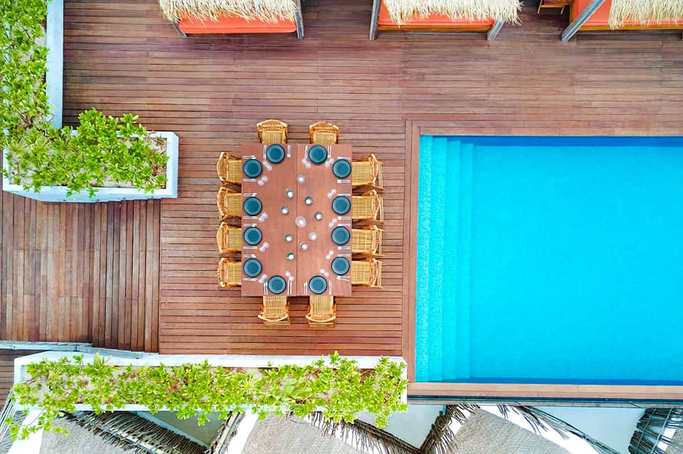 Aerial view of wooden deck with round dining table, pool, umbrellas, and tropical greenery