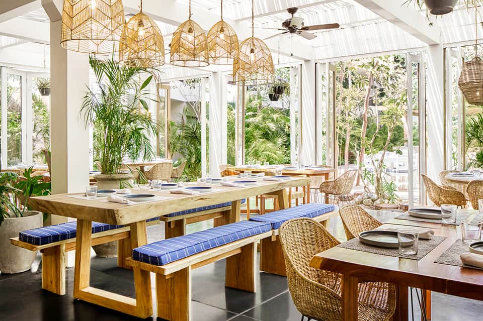Bright dining space with wooden table, wicker chairs, golden hanging lamps, and large windows overlooking gardens