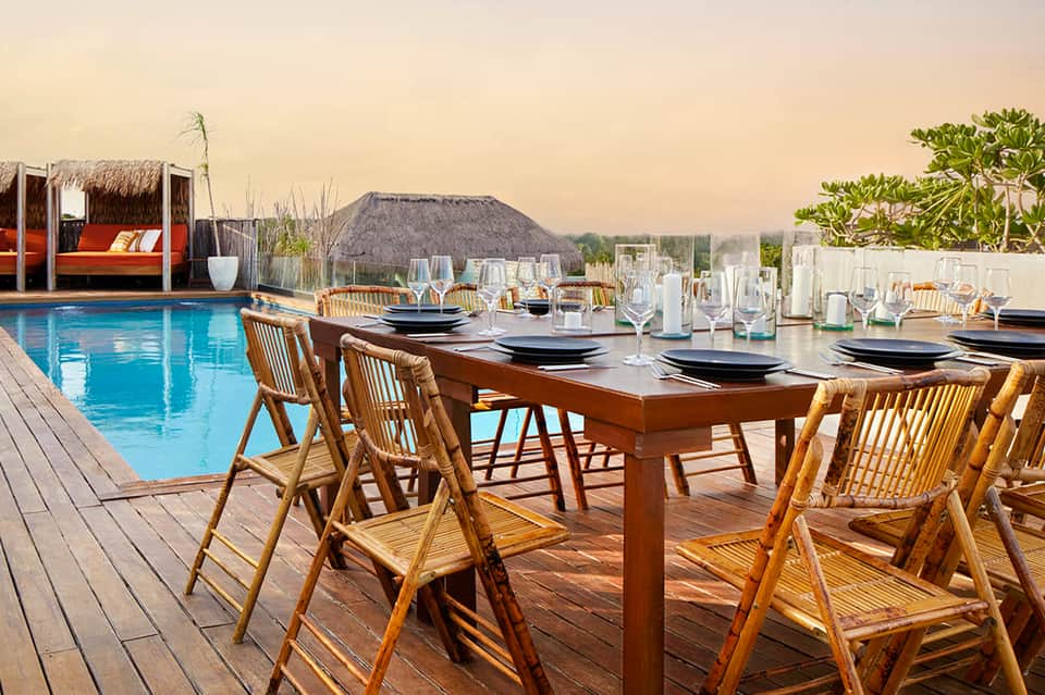 Rooftop dining setup with wooden chairs, table settings, pool, and thatched cabana at sunset