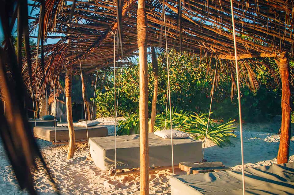 Covered sand area with swing beds under palm frond structures overlooking water