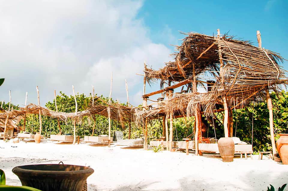 Tropical beach club with thatched-roof structures, white sand, wooden lounge chairs, and green vegetation