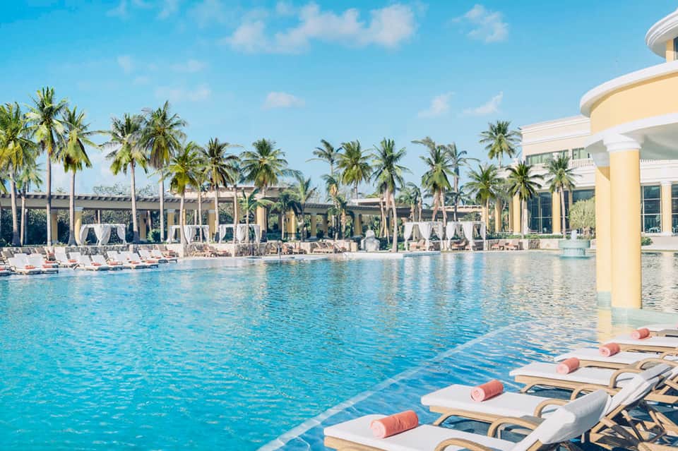 Resort lagoon pool surrounded by palm trees, lounge chairs, and cream-colored building architecture