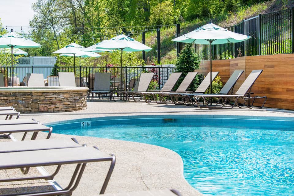 Resort swimming pool with lounge chairs, umbrellas, and stone fire pit surrounded by trees