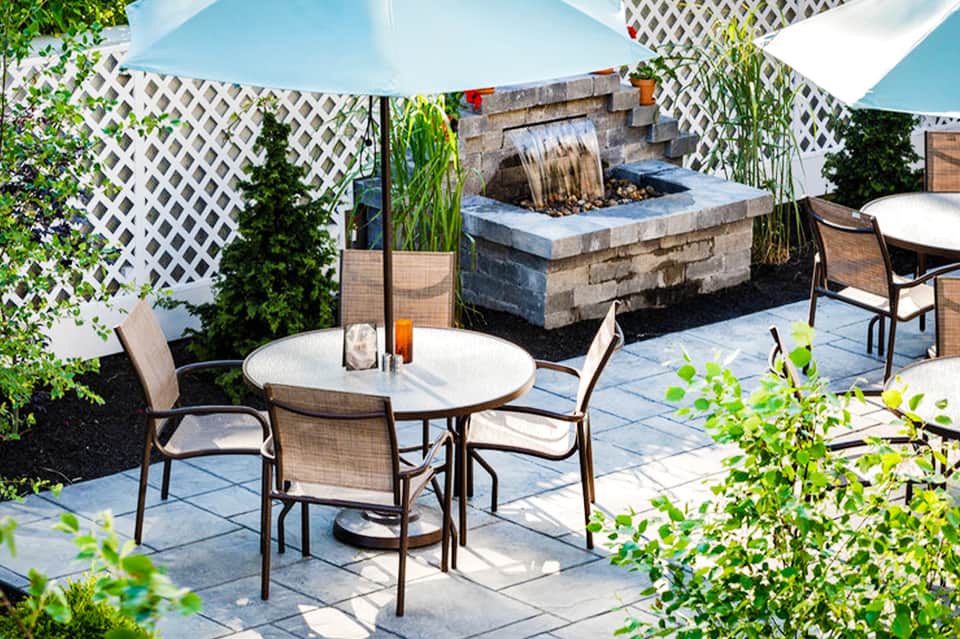 Upscale patio area with round dining table, white umbrellas, fire pit, white lattice trellis, and lush landscaping