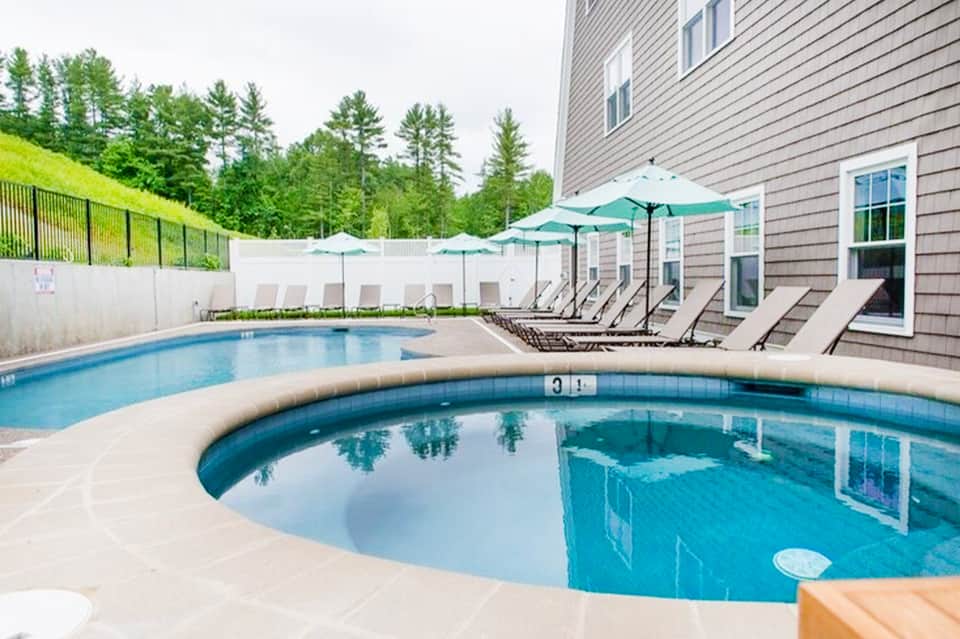 Residential pool area with hot tub, lounge chairs, and green umbrellas by house