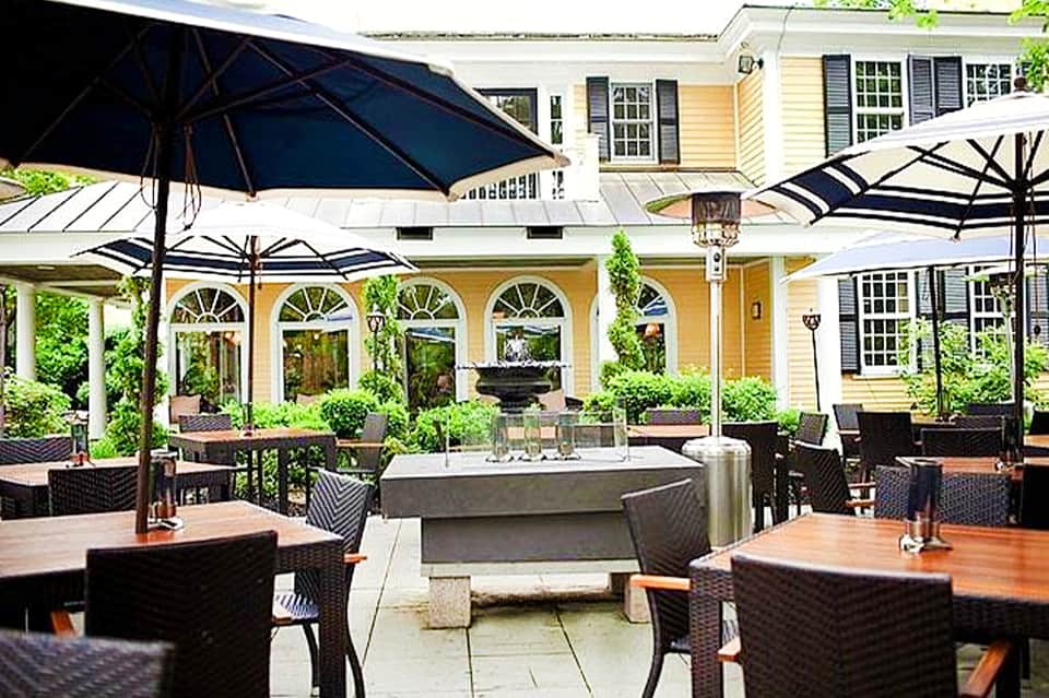 Outdoor patio dining area with black umbrellas, wooden tables, yellow architecture, and potted plants