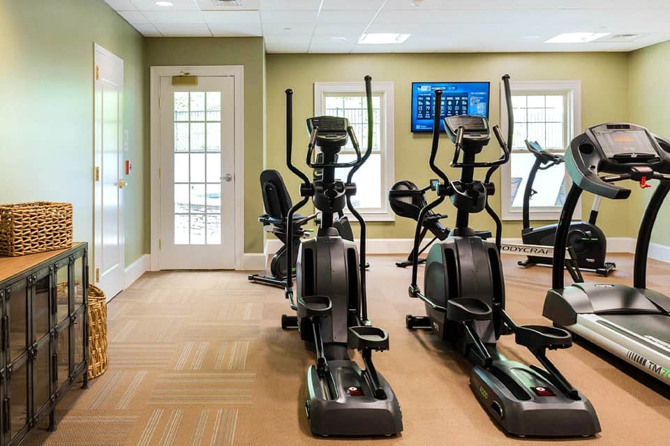 Bright fitness center with elliptical machines, treadmills, and large windows overlooking grounds