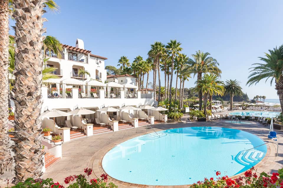 Beachfront pool area with white cabanas, lounge chairs, palm trees, and Mediterranean architecture